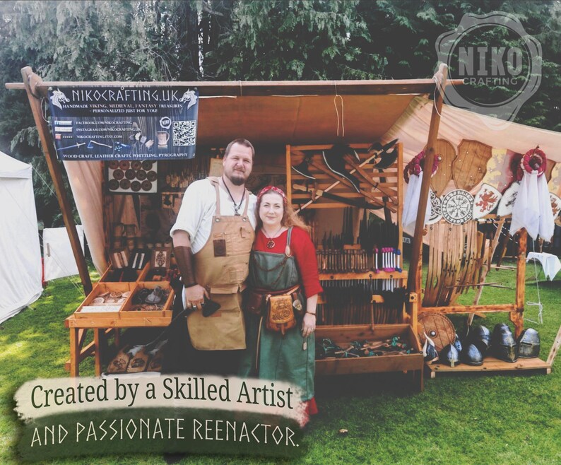 May include: A vendor booth displaying handmade Viking, medieval, and fantasy treasures. The booth features a variety of leather crafts, wooden items, and weaponry. The sign reads "NIKOCRAFTING.UK" and "Created by a Skilled Artist and Passionate Reenactor."