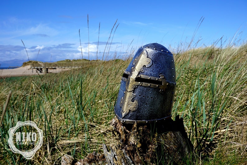 Op de afbeelding: Een zilveren en gouden ridderhelm met een kruisontwerp aan de voorkant. De helm staat op een boomstronk in een grasveld.