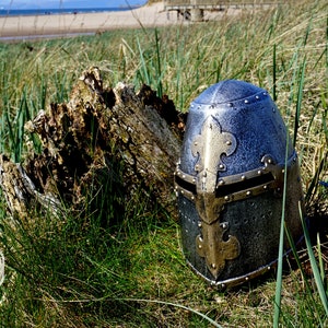 Op de afbeelding: Een zilveren en gouden ridderhelm met een fleur-de-lis-ontwerp aan de voorkant. De helm ligt op een bed van groen gras.
