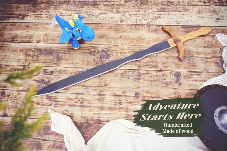 May include: A wooden sword with a gray blade and a light brown handle lies on a wooden surface. A blue plush dragon toy is nearby. The image includes the text "Adventure Starts Here".