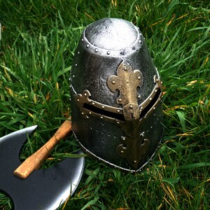 Op de afbeelding: Een zilveren en gouden ridderhelm met een fleur-de-lis-ontwerp ligt op een bed van groen gras. Een zwarte dubbelzijdige bijl met een houten handvat ligt naast de helm.