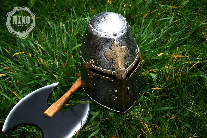 Op de afbeelding: Een zilveren en gouden ridderhelm met een fleur-de-lis-ontwerp ligt op een bed van groen gras. Een zwarte dubbelzijdige bijl met een houten handvat ligt naast de helm.