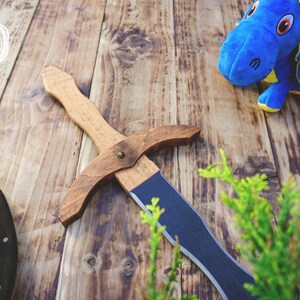 May include: A wooden toy sword with a brown handle and a blue blade, a blue plush dragon, and a black and blue shield are arranged on a wooden surface. The sword is approximately 18 inches long. The image has a rustic, playful theme.