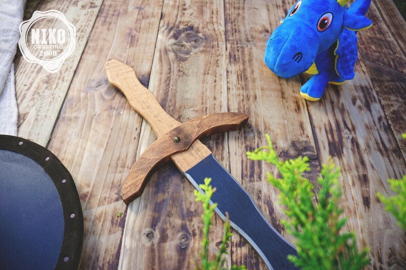 May include: A wooden toy sword with a brown handle and a blue blade, a blue plush dragon, and a black and blue shield are arranged on a wooden surface. The sword is approximately 18 inches long. The image has a rustic, playful theme.