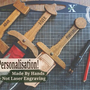May include: Wooden toy swords with personalized names, including "AIDAN", "RIVER", and "MARSHALL". The swords have leather sheaths and are displayed on a grid cutting mat. The image also includes text that says "Free Personalisation!" and "Made By Hands - Not Laser Engraving".