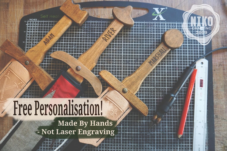 May include: Wooden toy swords with personalized names, including "AIDAN", "RIVER", and "MARSHALL". The swords have leather sheaths and are displayed on a grid cutting mat. The image also includes text that says "Free Personalisation!" and "Made By Hands - Not Laser Engraving".