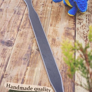 May include: A handmade wooden sword with a gray blade and a brown handle rests on a wooden surface. A blue plush dragon toy with yellow accents is nearby. The text "Handmade quality, Natural materials, Built to last" is visible.
