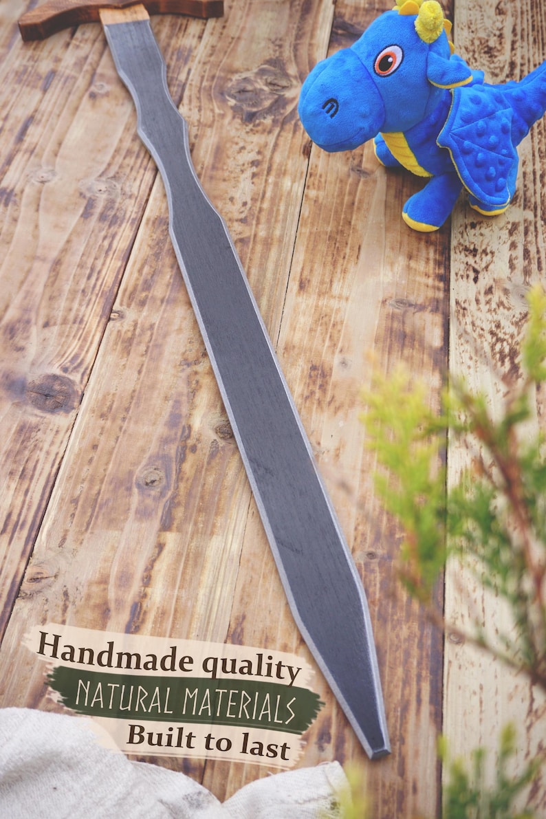May include: A handmade wooden sword with a gray blade and a brown handle rests on a wooden surface. A blue plush dragon toy with yellow accents is nearby. The text "Handmade quality, Natural materials, Built to last" is visible.