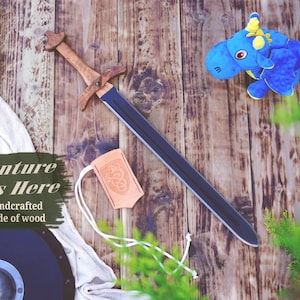 May include: A wooden toy sword with a dark blade and light brown hilt, a small tan leather pouch with a dragon emblem, a blue plush dragon toy, and a round dark shield. Text on the image reads: Adventure Starts Here Handcrafted Made of wood.