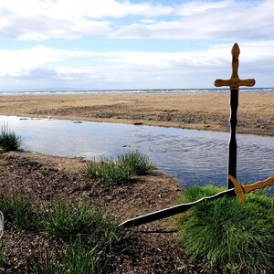 May include: Two wooden swords, one upright and one lying on the ground, are positioned on a grassy area near a stream. The swords have gold-colored handles and dark blades. The background features a sandy beach and a cloudy sky.