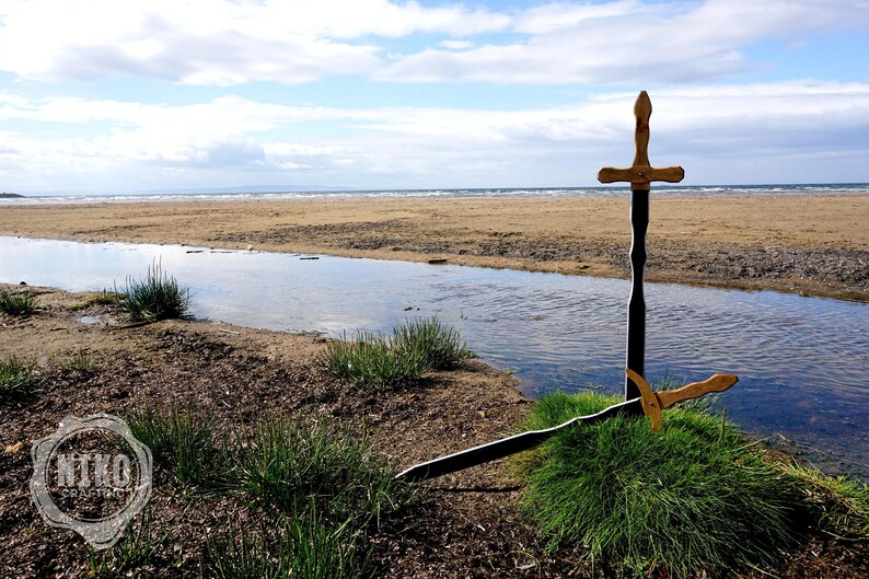 May include: Two wooden swords, one upright and one lying on the ground, are positioned on a grassy area near a stream. The swords have gold-colored handles and dark blades. The background features a sandy beach and a cloudy sky.