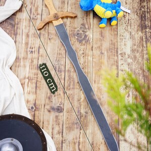 May include: A wooden toy sword, approximately 43 inches long, with a light brown handle and crossguard, and a gray blade. A blue and yellow stuffed dragon toy is also present, along with a black shield and white cloth.