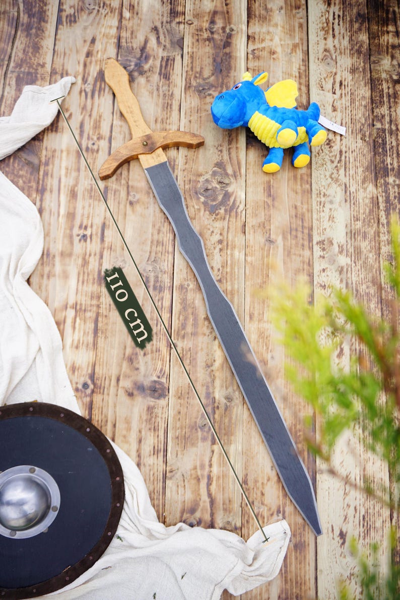 May include: A wooden toy sword, approximately 43 inches long, with a light brown handle and crossguard, and a gray blade. A blue and yellow stuffed dragon toy is also present, along with a black shield and white cloth.