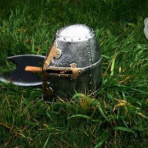 Op de afbeelding: Een zilveren en gouden ridderhelm met een zwarte bijl die op het gras ligt.