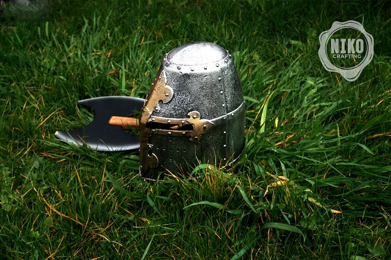 Op de afbeelding: Een zilveren en gouden ridderhelm met een zwarte bijl die op het gras ligt.