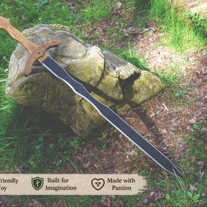 May include: A wooden toy sword with a light brown handle and crossguard, and a black blade. The sword rests on a moss-covered rock in a natural outdoor setting. The image includes text that reads "Eco-Friendly Toy", "Built for Imagination", and "Made with Passion".