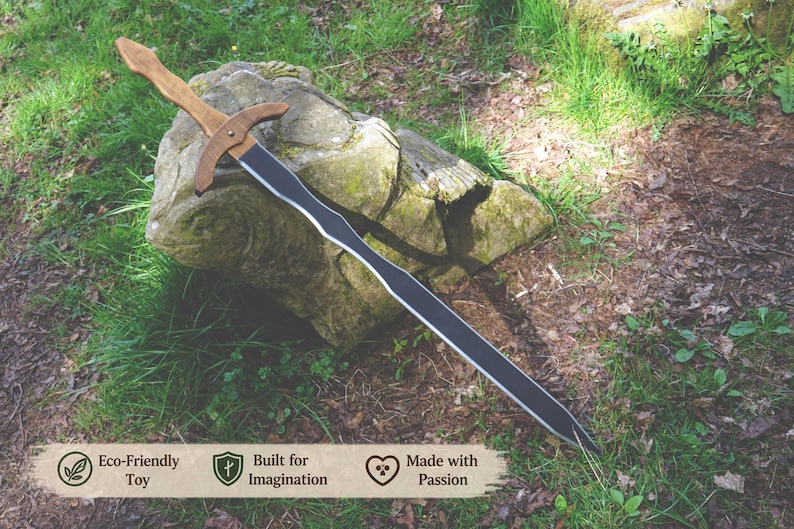May include: A wooden toy sword with a light brown handle and crossguard, and a black blade. The sword rests on a moss-covered rock in a natural outdoor setting. The image includes text that reads "Eco-Friendly Toy", "Built for Imagination", and "Made with Passion".