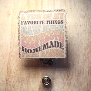 May include: A rectangular badge reel with a glittery border. The badge reel has the text "A FEW OF MY FAVORITE THINGS" and lists "RAW MILK, SOURDOUGH, BONE BROTH, HOMEMADE, FREE RANGE" in a retro font. The reel has a retractable cord and a snap.