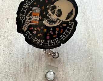 May include: A black badge reel featuring the text "SLINGIN' PILLS TO PAY THE BILLS" and a skull graphic holding a pill bottle. The reel has a retractable cord and a clear plastic snap.