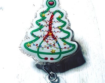 May include: A Christmas tree-shaped badge reel with a glittery border. The tree is white with green outlines and a red center. It is decorated with colorful lights and gold stars. A heart is at the top of the tree.