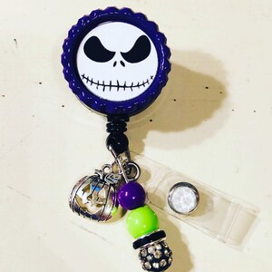May include: A badge reel with a design of Jack Skellington's face from The Nightmare Before Christmas. The purple reel has a scalloped edge. A charm of a pumpkin, purple and green beads, and a rhinestone bead dangle from the reel.