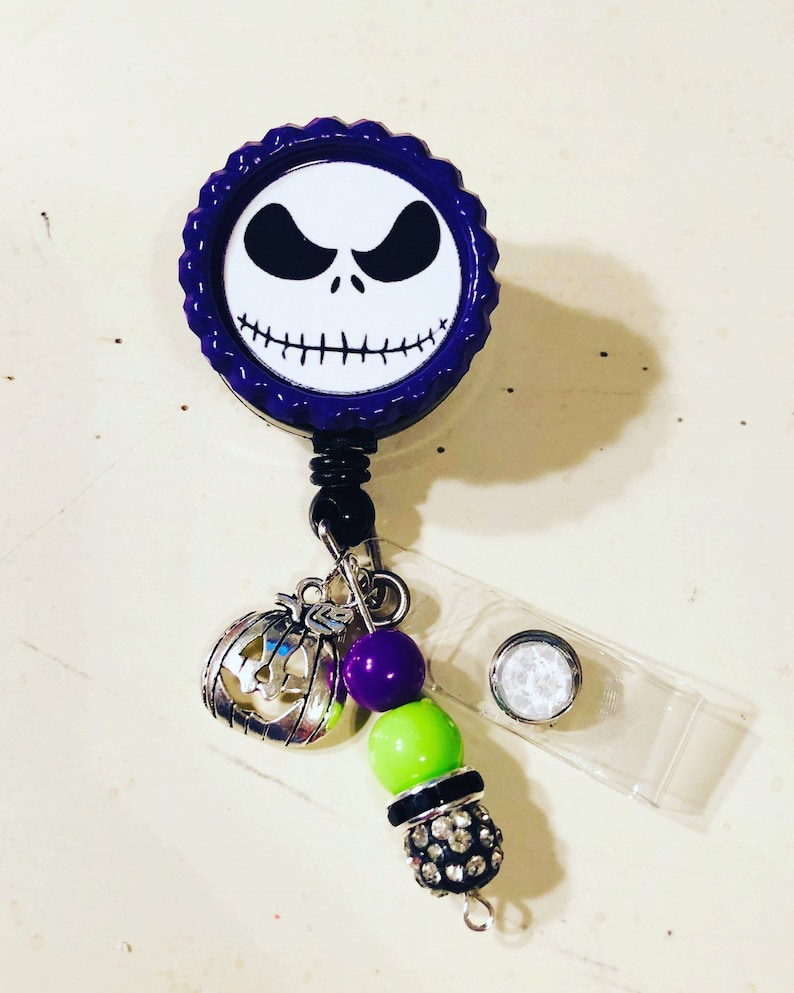 May include: A badge reel with a design of Jack Skellington's face from The Nightmare Before Christmas. The purple reel has a scalloped edge. A charm of a pumpkin, purple and green beads, and a rhinestone bead dangle from the reel.