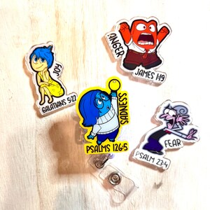May include: A collection of five acrylic badge reels featuring characters from the movie "Inside Out." The characters include Joy, Anger, Sadness, and Fear, each with associated text. The reels are clear with a metal clip.