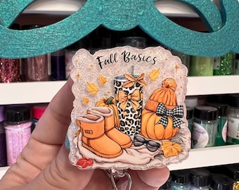May include: A badge reel with a fall theme. The design includes orange boots, a leopard print tumbler, a pumpkin with a hat, and the text "Fall Basics". The background is glittery with autumn leaves.