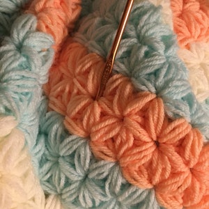 May include: Close-up of a crocheted blanket with a repeating pattern of flowers in white, light blue, and peach. A gold crochet hook is resting on the blanket. The hook is labeled "Size 7-8 US".