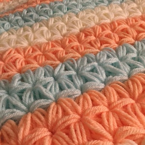 May include: A close-up of a crocheted blanket with stripes of peach, light blue, and white yarn. The blanket is made with a textured stitch that creates a raised pattern.