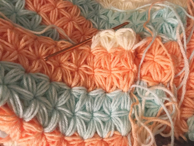 May include: A close-up of a crocheted blanket with a repeating pattern of flowers in shades of peach, light blue, and white. A crochet hook is visible in the image.
