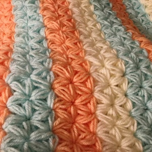 May include: A crocheted baby blanket with stripes of peach, light blue, and white yarn. The blanket is made with a textured stitch that creates a raised pattern.