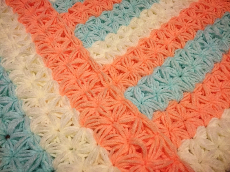 May include: A crocheted blanket with stripes of peach, white and light blue. The blanket is made with a textured stitch that creates a flower-like pattern.