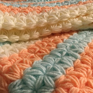 May include: A crocheted baby blanket with stripes of white, peach, and light blue yarn. The blanket is made with a textured stitch that creates a raised, puffy effect.