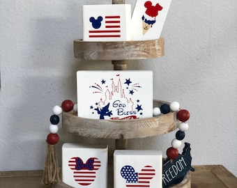 May include: A three-tiered wooden tray with a red, white, and blue theme. The top tier has a red, white, and blue ice cream cone with Mickey Mouse ears. The middle tier has a white square with a red, white, and blue American flag and Mickey Mouse ears. The bottom tier has a white square with a red, white, and blue American flag and Mickey Mouse ears, a white square with a red, white, and blue American flag and Minnie Mouse ears, and a blue and white wooden cutout of the United States with the word "FREEDOM" written on it. The tray is decorated with a red, white, and blue beaded garland.