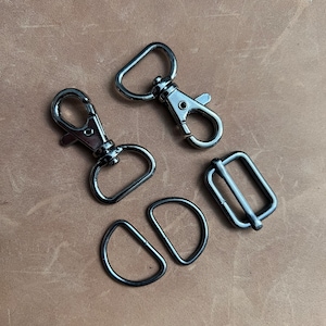 May include: A collection of silver-toned metal hardware components, including swivel clasps, D-rings, and a rectangular slider buckle. The items are arranged on a light brown, textured surface, likely leather or a similar material. These components are commonly used in bag making and crafting.