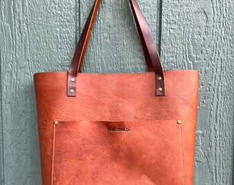 May include: A brown leather tote bag with a front pocket and brass rivets. The bag has a brown leather strap and the text "Pacific Stitch Co." is embossed on the front pocket.