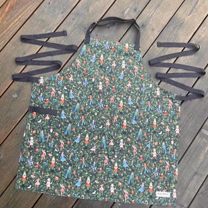 May include: A green apron with a festive pattern of nutcrackers, ballerinas, and candy canes. The apron has black straps and a pocket. The apron is made of fabric and is designed for kitchen use.