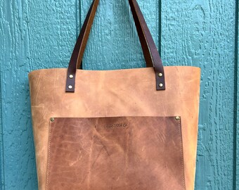 May include: A tan leather tote bag with a brown leather pocket and handles. The bag has a rectangular pocket on the front with the text "Celtic Stitch Co." The handles are attached with brass rivets. The bag is hanging against a blue wooden background.