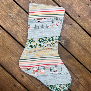 May include: Quilted Christmas stocking with a variety of patterns and colors. The stocking features stripes, winter scenes with houses and trees, and floral designs. The top has a loop for hanging. The background is a wooden surface.