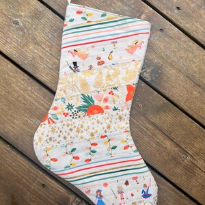 May include: A white Christmas stocking with a patchwork design featuring a variety of holiday-themed patterns, including stripes, stars, and illustrations of angels, nutcrackers, and other festive figures.