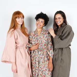 May include: Three women wearing different robes. The woman on the left is wearing a pink robe, the woman in the middle is wearing a floral robe, and the woman on the right is wearing a dark green robe.