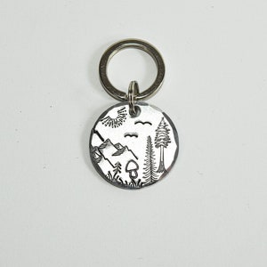 Può includere: Un portachiavi d'argento con un ciondolo rotondo che presenta un designo inciso di una catena montuosa, un sole, un fungo, due alberi e due uccelli in volo.