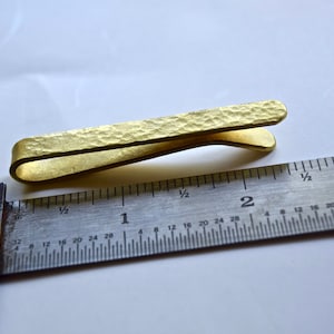 Op de afbeelding: Een gouden metalen stropdasclip met een gehamerde textuur, gemeten met een liniaal. De stropdasclip is ongeveer 3,8 cm lang.