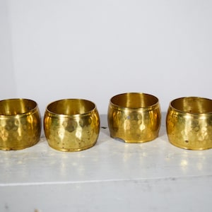 May include: Four gold napkin rings with a hammered texture. The rings are cylindrical and sit on a white surface. The napkin rings are made of metal and have a shiny finish.