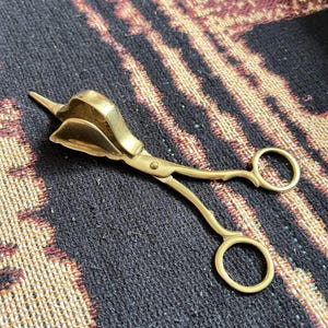 May include: A brass candle snuffer with a scissor-like design. The snuffer has a cup-shaped head to extinguish candles and a pointed tip. The handles have circular finger holes. The object is on a dark patterned surface.