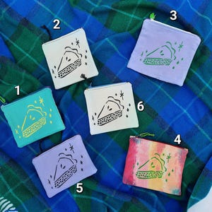 May include: A collection of six small, square zipper pouches in various colors, each featuring a pie slice graphic. The pouches are teal, white, lavender, and a watercolor-style design. The background is a blue and green plaid blanket.