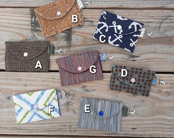 May include: A selection of small fabric wallets in various patterns and colours. Designs include brown corduroy, navy with white anchors, and blue and green plaid. Each wallet has a snap closure and a key ring attachment.