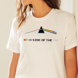 Pu&ograve; includere: Maglietta bianca con un triangolo nero, un arcobaleno e un gatto rosa. Il testo "NYAN SIDE OF THE CAT" &egrave; stampato sotto la grafica.