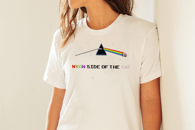 Pu&ograve; includere: Maglietta bianca con un triangolo nero, un arcobaleno e un gatto rosa. Il testo "NYAN SIDE OF THE CAT" &egrave; stampato sotto la grafica.